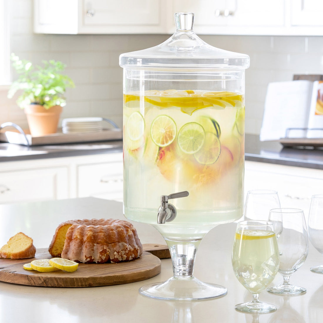 Large glass drink dispenser with lemon slices and a bundt cake on a kitchen counter.