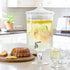 Large glass drink dispenser with lemon slices and a bundt cake on a kitchen counter.