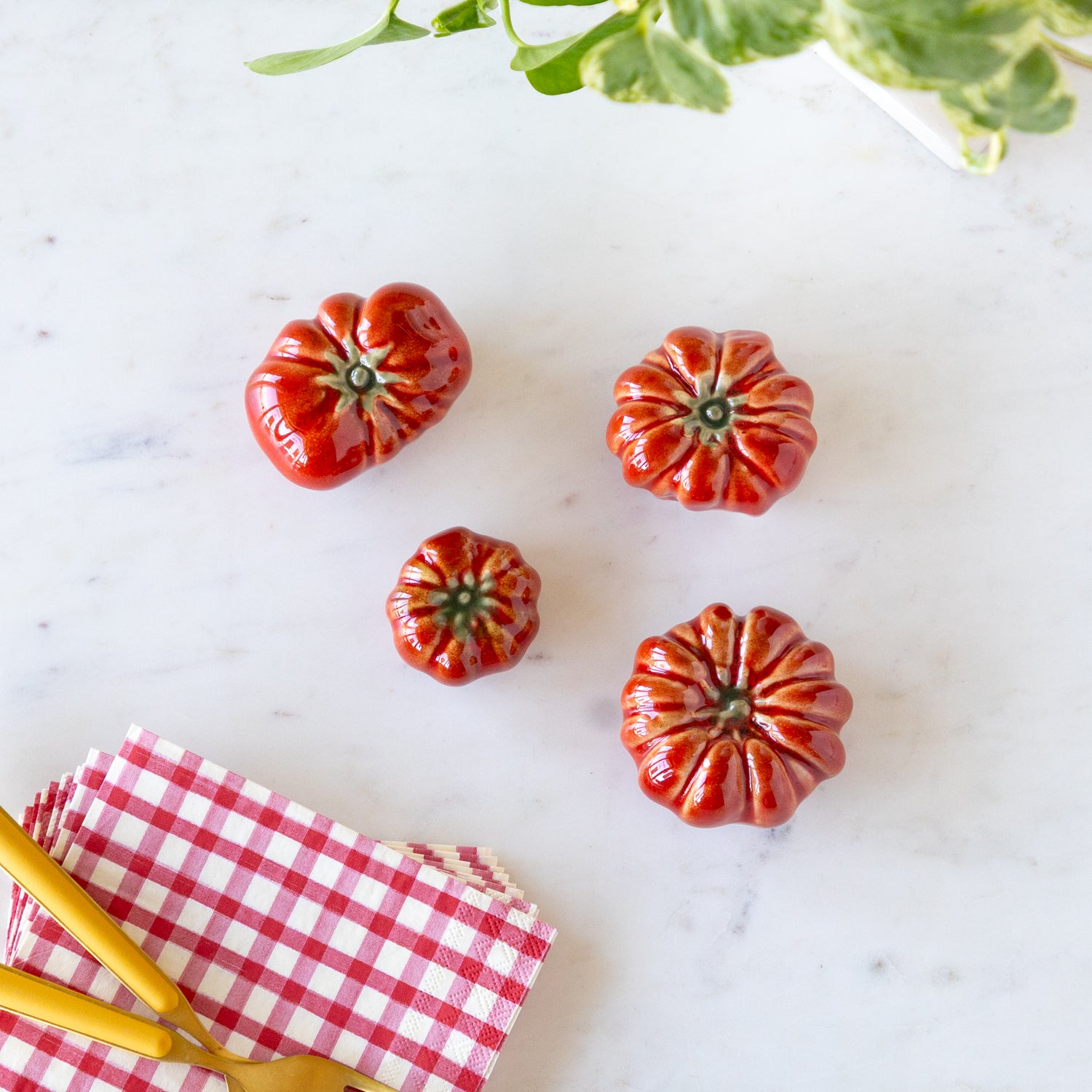 Red ceramic tomatoes on a marble surface with gold forks and a red checkered napkin.