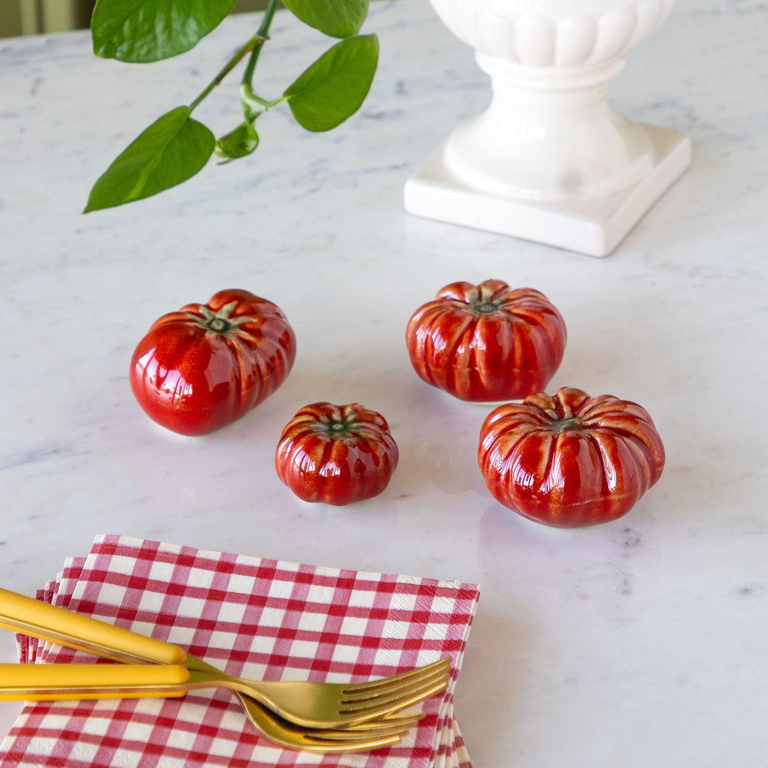 Red ceramic tomatoes on a marble surface with gold forks and a red checkered napkin.