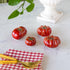 Red ceramic tomatoes on a marble surface with gold forks and a red checkered napkin.