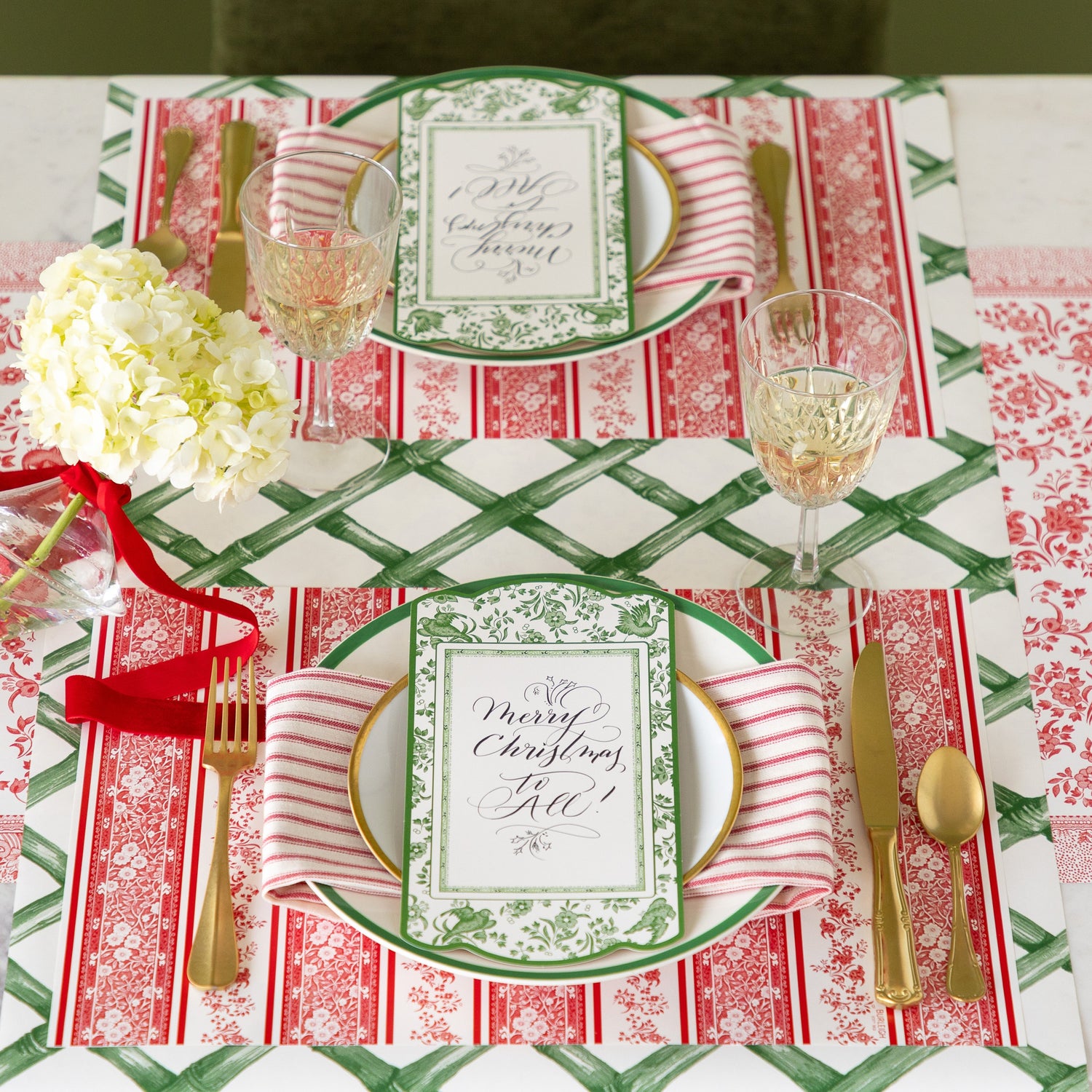 A table setting featuring the Red Burleigh Stripe Placemat, green rimmed dinner and gold rimmed salad plates with a red stripe napkin and Burleigh Green Regal Peacock Table Accent reading &quot;Merry Christmas to All!&quot; atop, gold flatware, glass of wine and Green Lattice Runner and Red Burleigh Runner underneath.