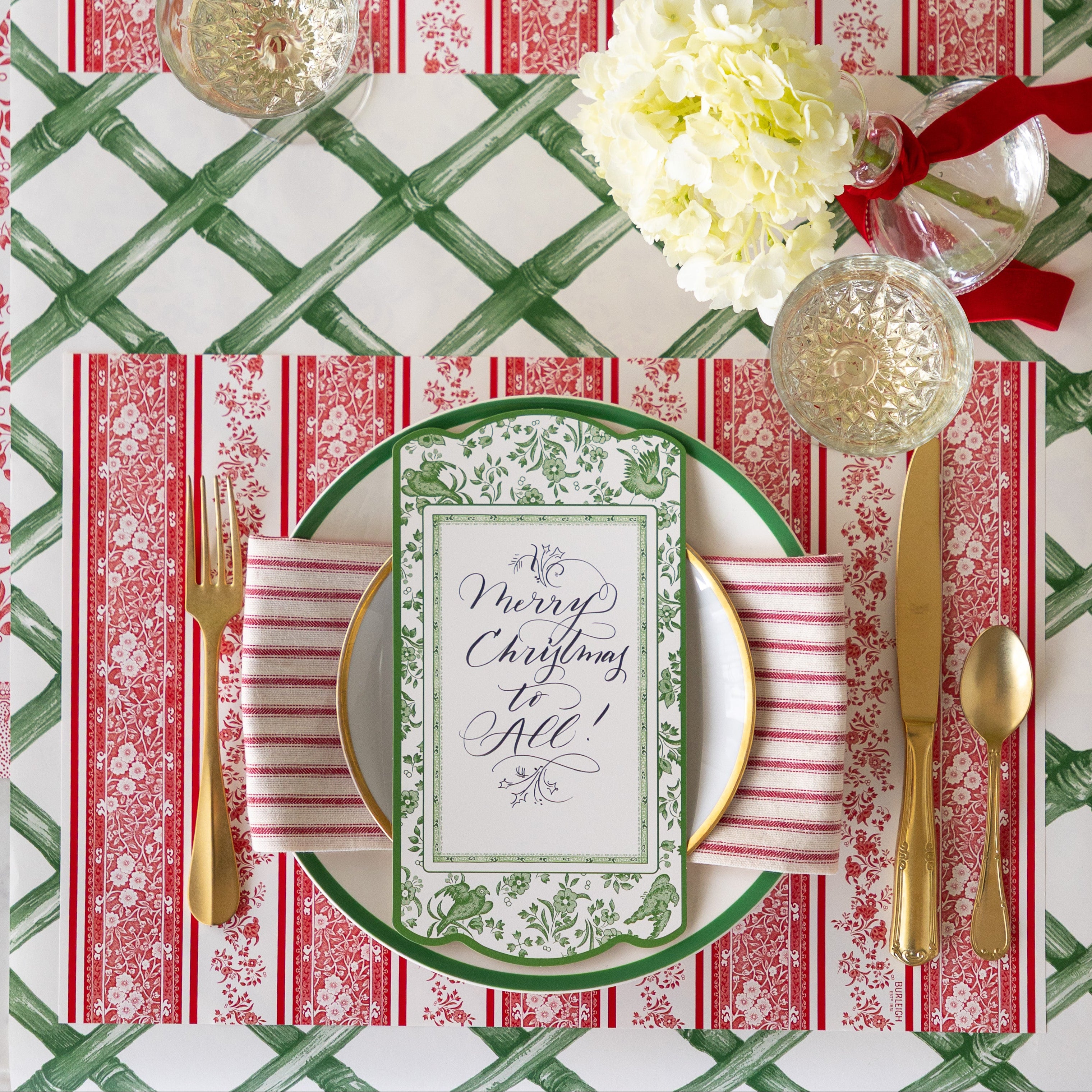 A place setting featuring the Red Burleigh Stripe Placemat, green rimmed and gold rimmed dinner plates with a red stripe napkin and Burleigh Green Regal Peacock Table Accent reading &quot;Merry Christmas to All!&quot; atop, gold flatware, glass of wine and Green Lattice Runner underneath.
