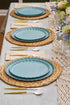 Table setting with blue plates, gold cutlery, and woven placemats on a light gray tablecloth.
