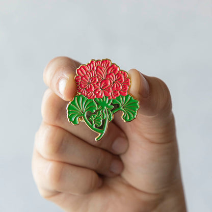 A person proudly displaying a Hester &amp; Cook Geranium Enamel Pin with a vibrant red chrysanthemum flower.