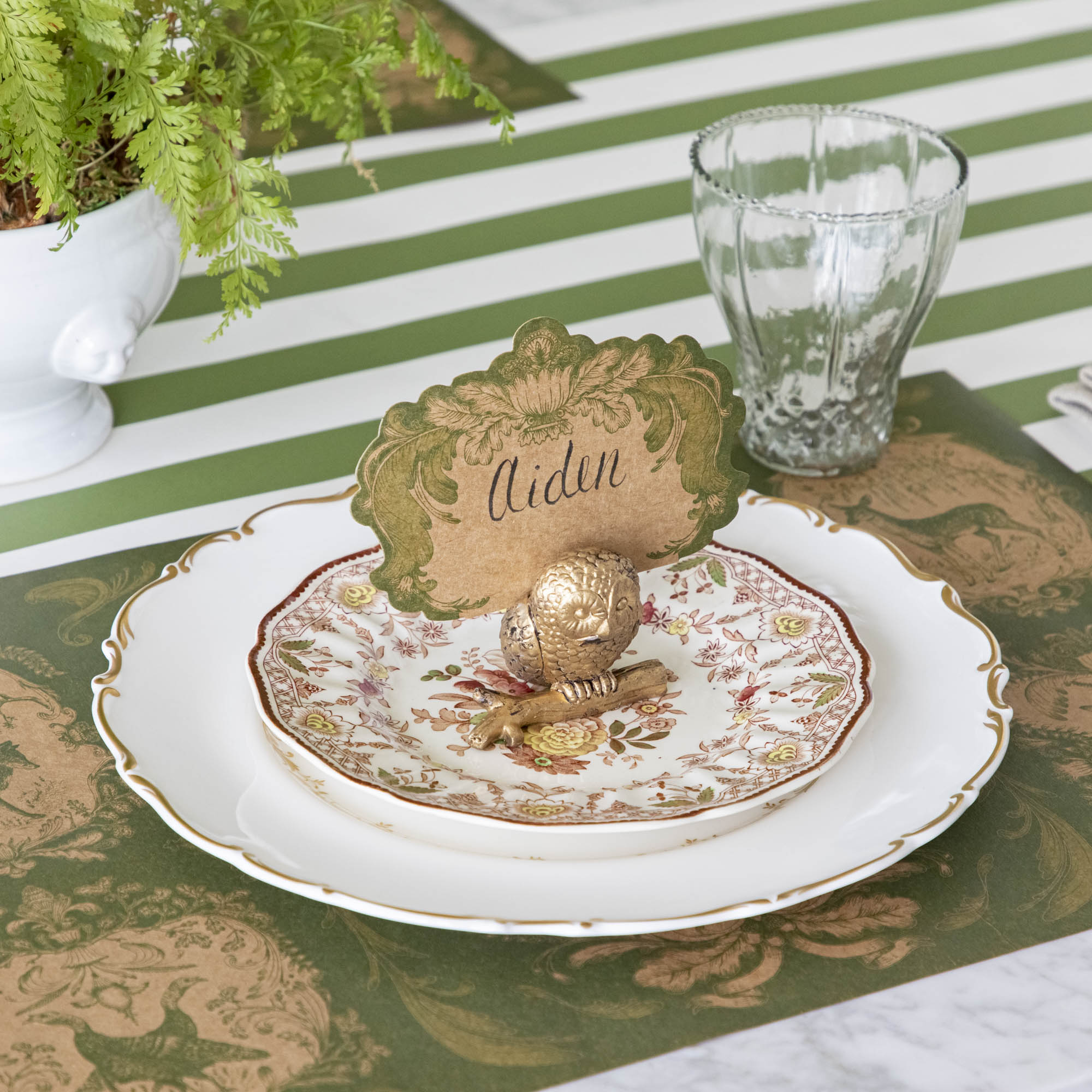 The antique gold finish Owl Name Card Holder, holding the Moss Fable Toile Place Card with &quot;Aiden&quot; written on it, on a table setting with two dinner plates, the Moss Fable Toile Placemat and Moss Green Classic Stripe Runner underneath.