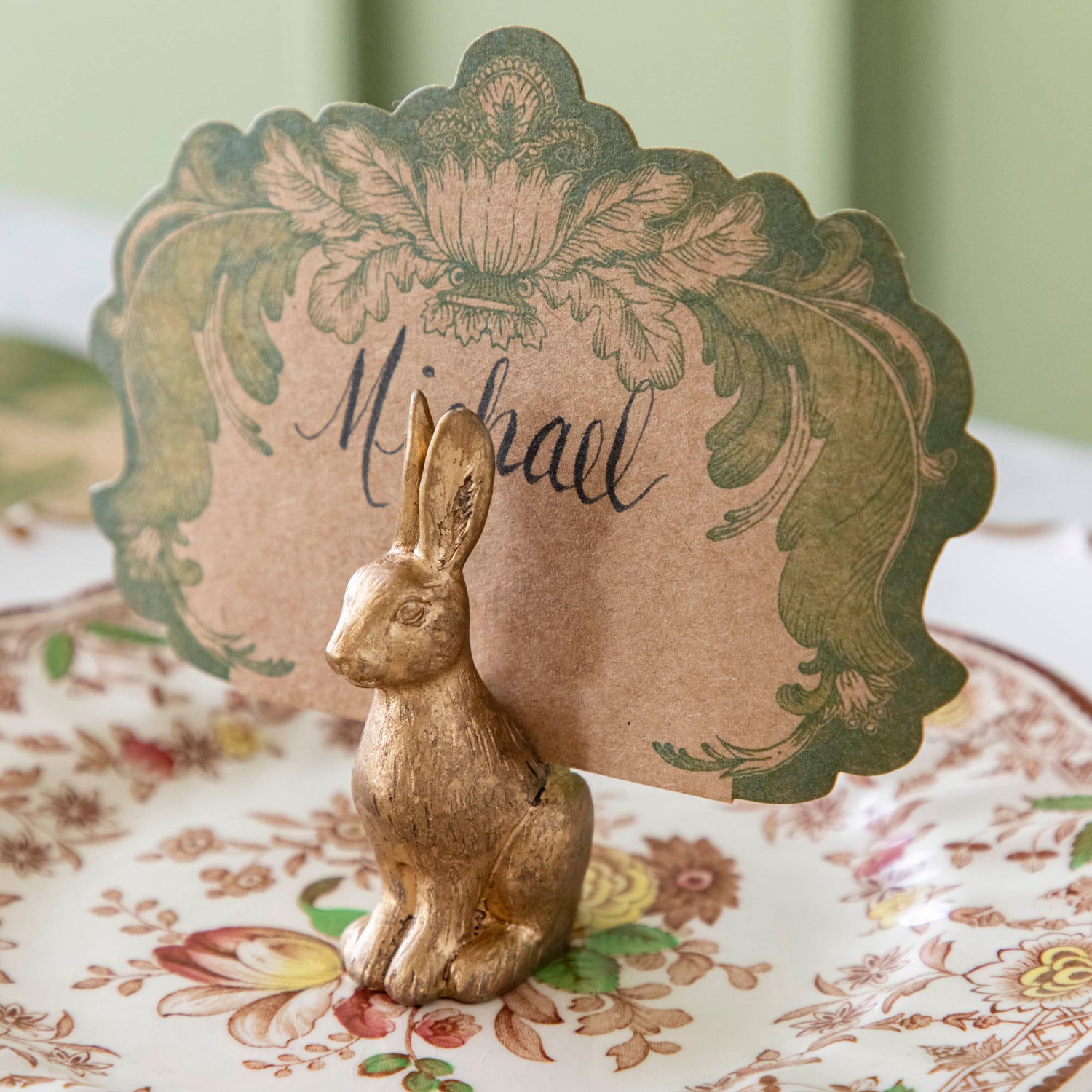 The antique gold finish Rabbit Name Card holder, holding the Moss Fable Toile Place Card with &quot;Michael&quot; written in cursive, sitting on a floral plate.