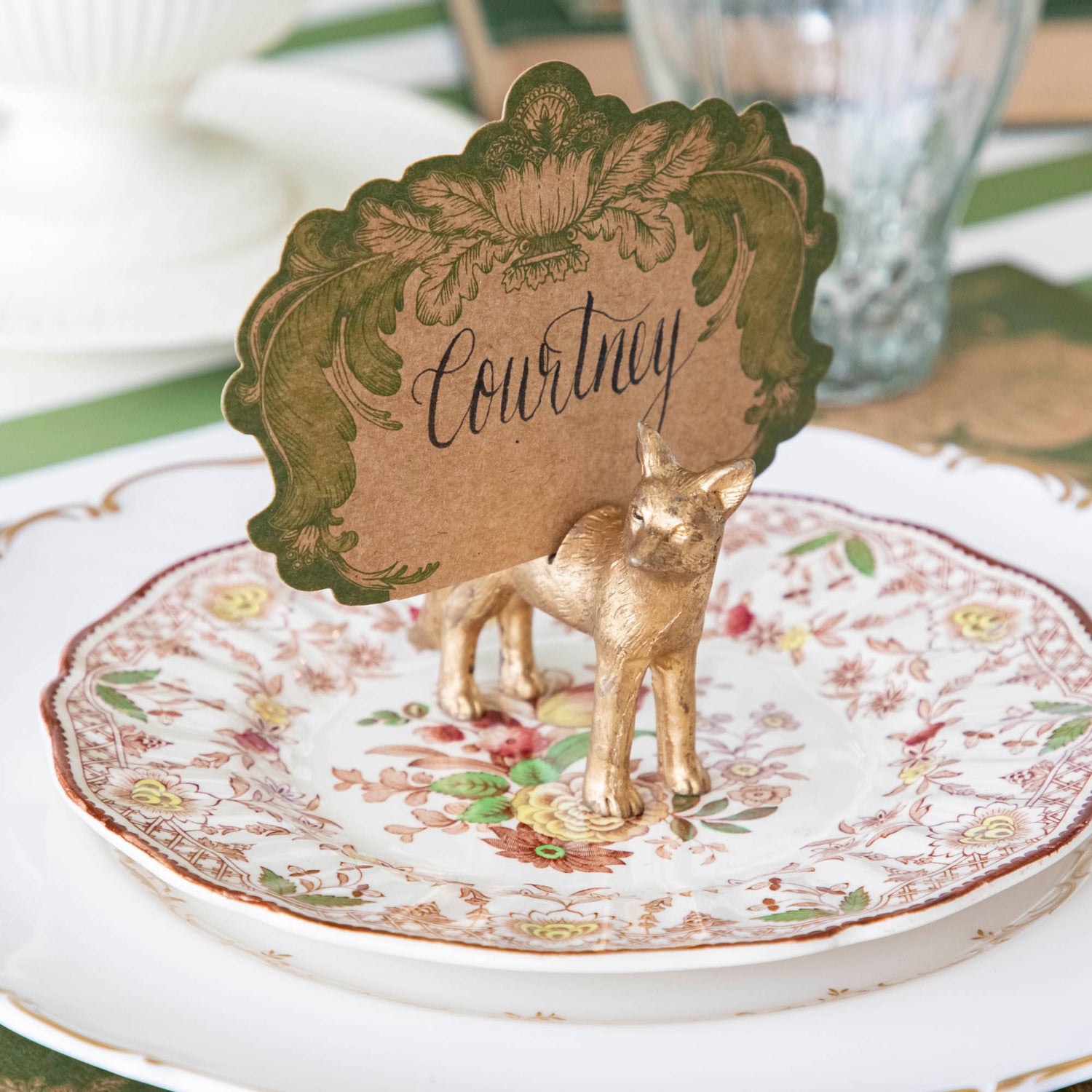 An antique gold finish Fox Name Card Holder, holding the Moss Fable Toile Place Card with &quot;Courtney&quot; written on it, standing on a floral plate with a white, gold trimmed plate underneath.