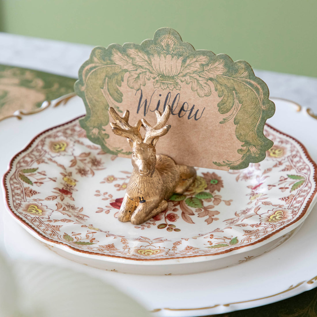 An antique gold finish Deer Place Card Holder on a floral dinner plate, holding the Moss Fable Toile Place Card with the name &quot;Willow&quot; written on it in cursive.