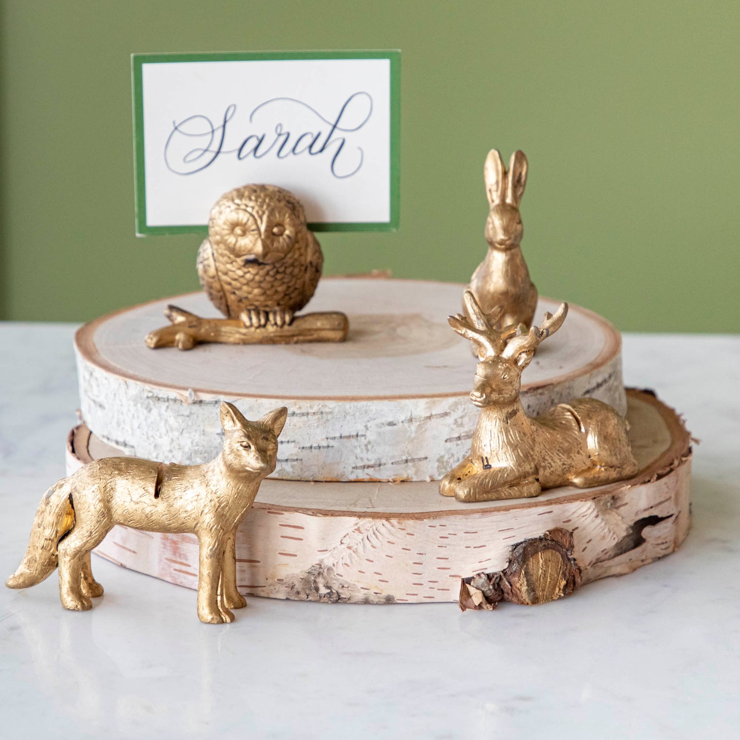 An Owl, Rabbit, Fox and Deer place card holder stacked on cylinders of wood with the Dark Green Frame Place Card with &quot;Sarah&quot; written on it in cursive, being held up by a slot in the owl&