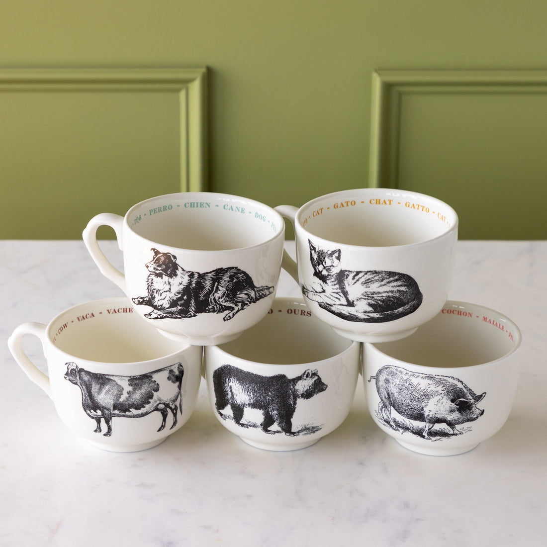 Set of five white mugs with black animal illustrations on a marble surface with a green wall background.