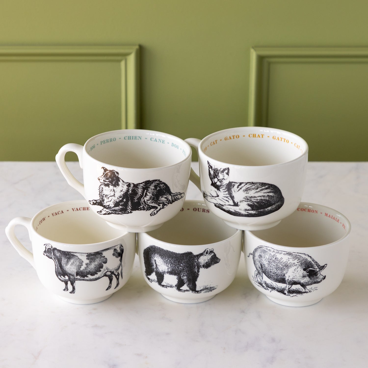 Set of five white mugs with black animal illustrations on a marble surface with a green wall background.