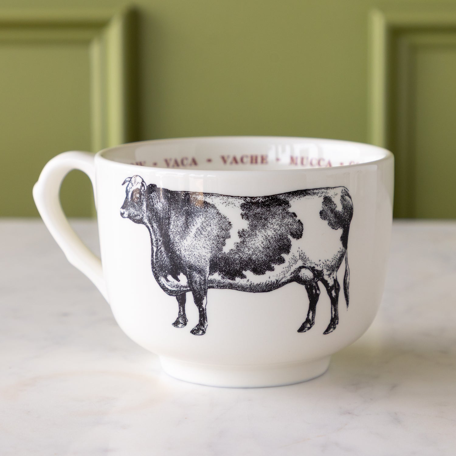 White mug with black cow illustration with &quot;cow&quot; in different languages in red text around the inside rim.