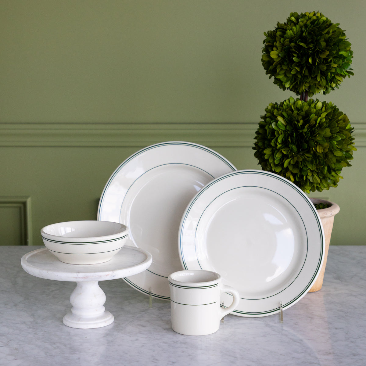 A set of white Fishs Eddy HL Greenband Dinnerware, including a mug, two plates, and a bowl on a white marble pedestal, styled on a marble counter against a green paneled wall next to a potted topiary.