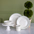 A set of white Fishs Eddy HL Greenband Dinnerware, including a mug, two plates, and a bowl on a white marble pedestal, styled on a marble counter against a green paneled wall next to a potted topiary.