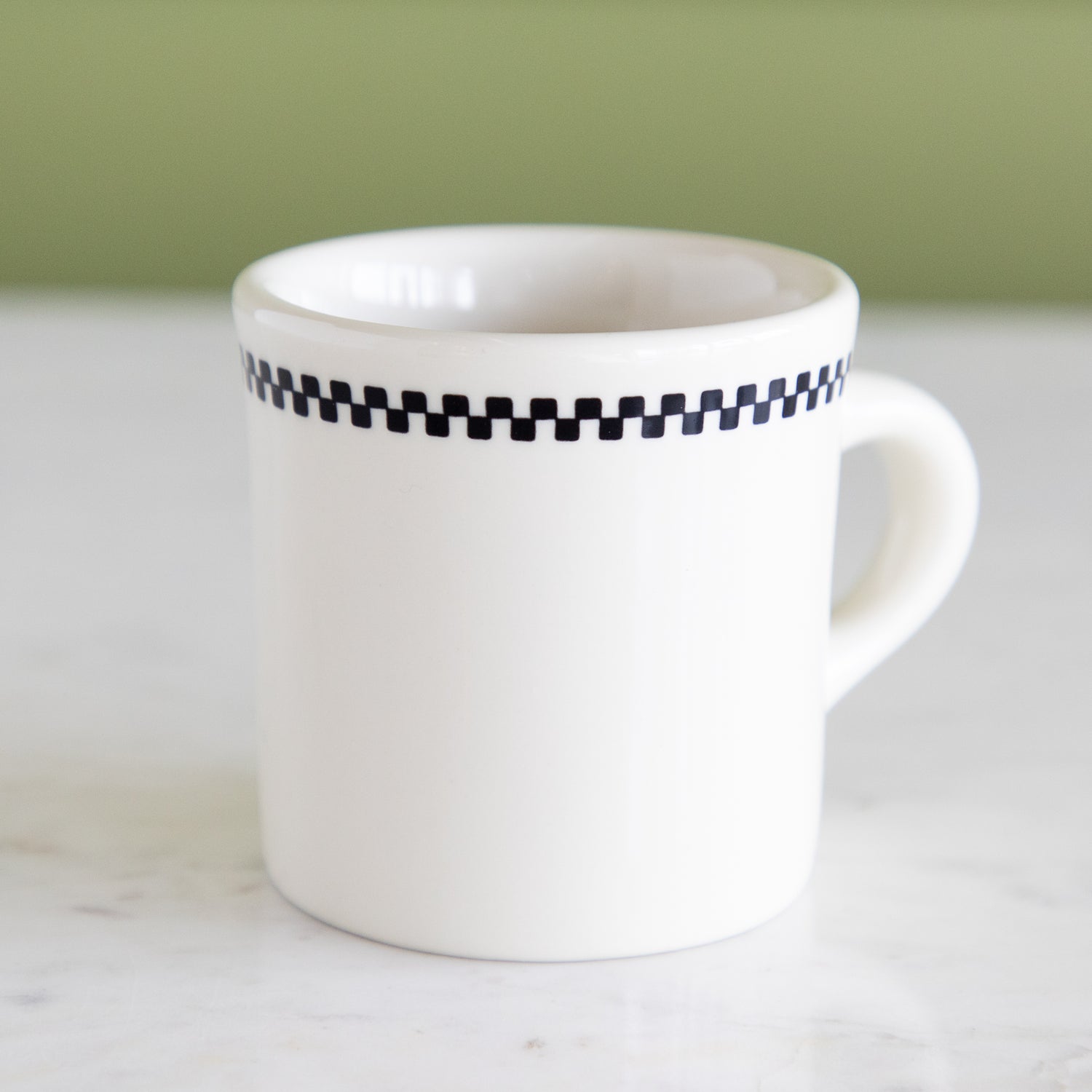 White mug with a black checkered pattern on a white surface with a green background.
