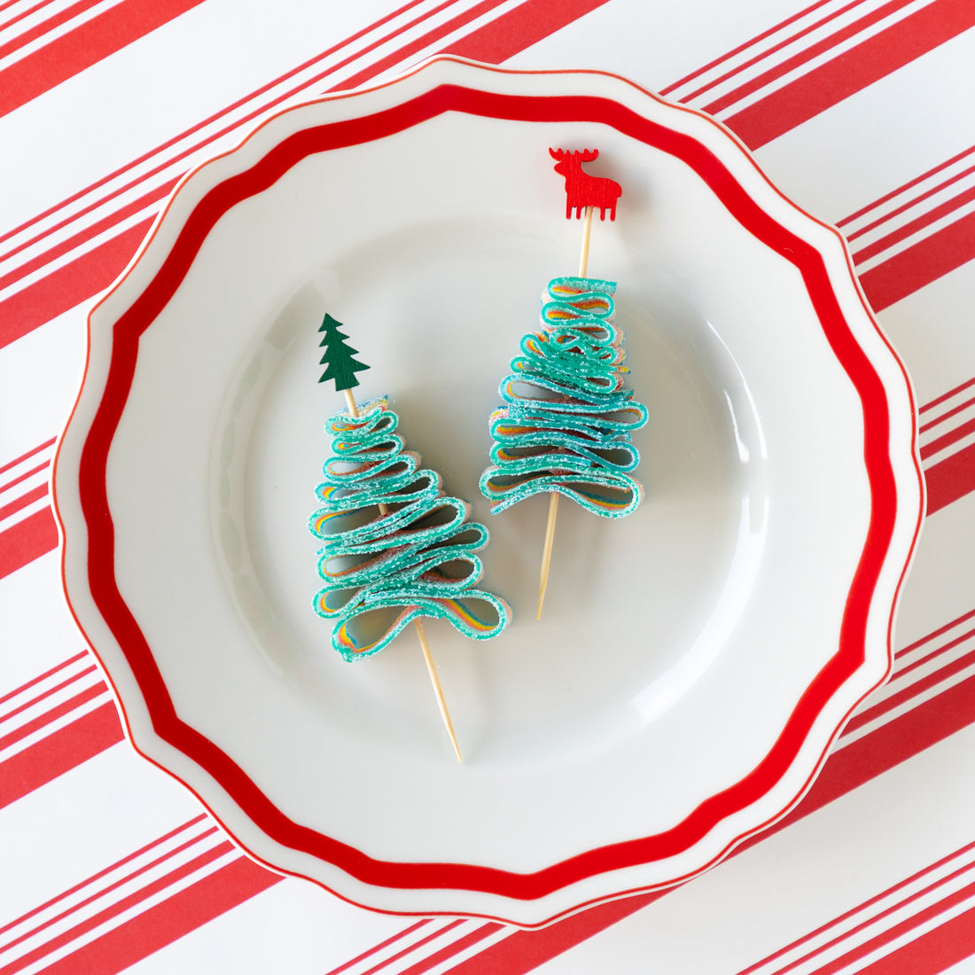 Deer and tree bamboo skewers piercing a candy rainbow rope folded in the shape of a tree, on a decorative plate and Peppermint Stripe Runner underneath.