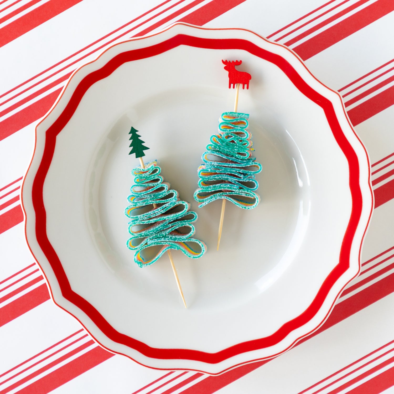Deer and tree bamboo skewers piercing a candy rainbow rope folded in the shape of a tree, on a decorative plate and Peppermint Stripe Runner underneath.