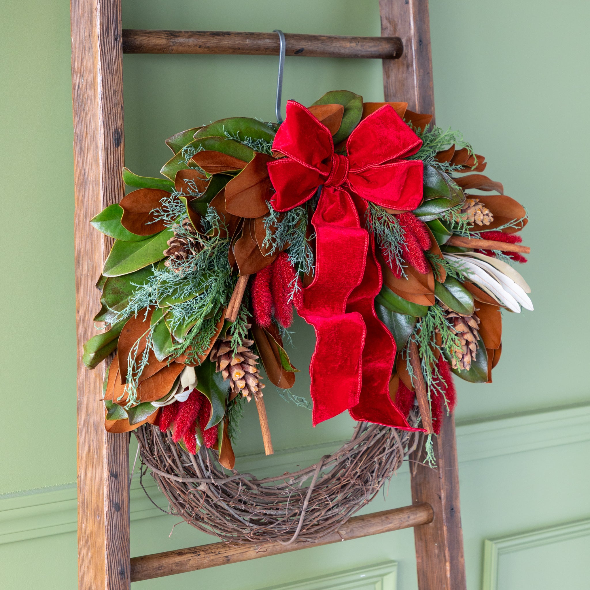 Decorative wreath with red bows and greenery on a wooden ladder against a green wall.