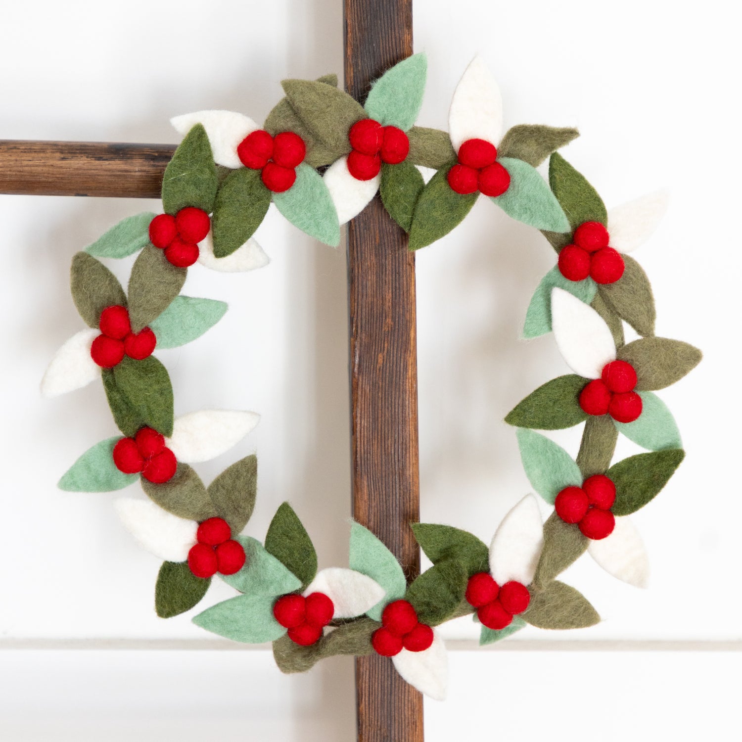Decorative felt wreath with red berries and green leaves on a wooden ladder against a white background.