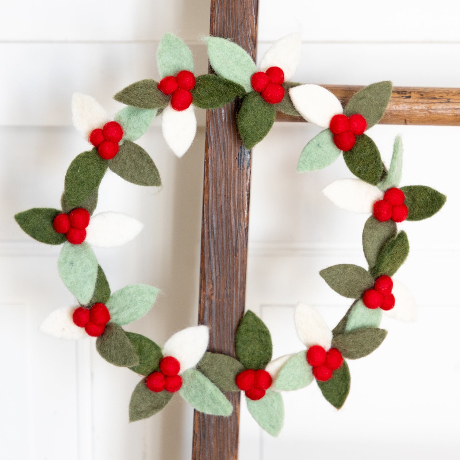 Decorative felt wreath with red berries and green leaves on a wooden ladder against a white background.