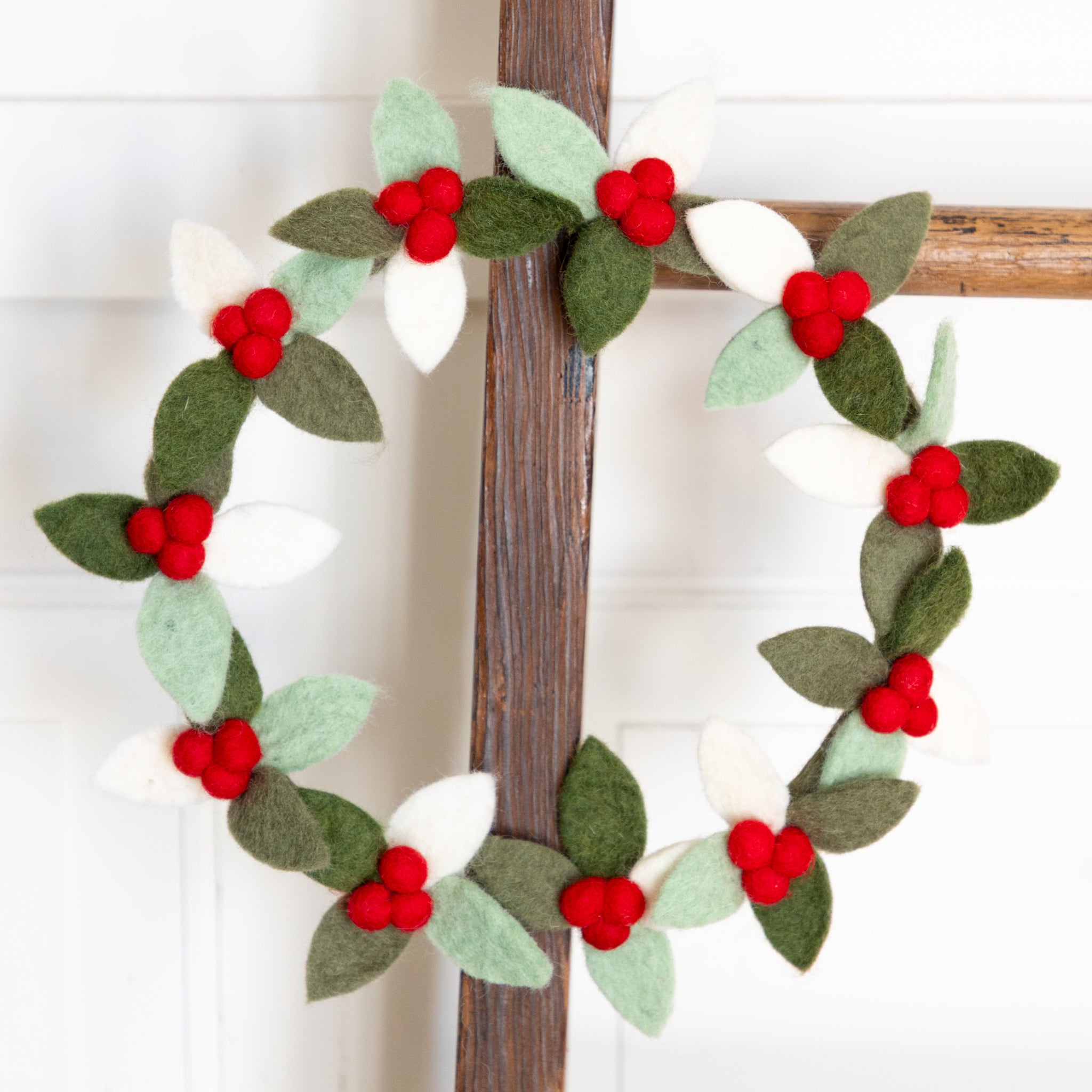 Decorative felt wreath with red berries and green leaves on a wooden ladder against a white background.