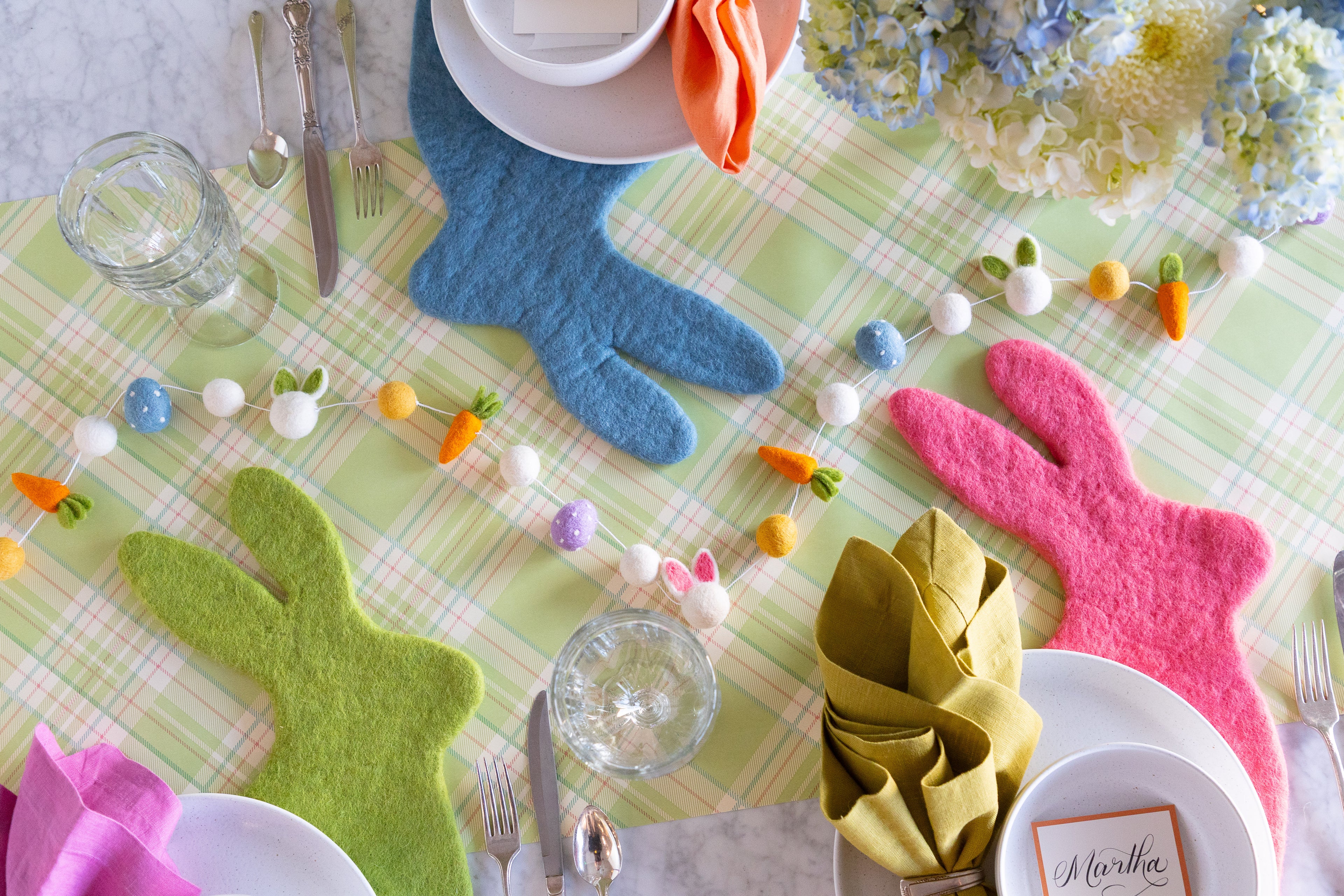 Easter themed table setting featuring flowers, Easter Felt Garland, Cornflower Blue, Spring Green and Pink Pom Felt Placemats on the Green Plaid Runner.