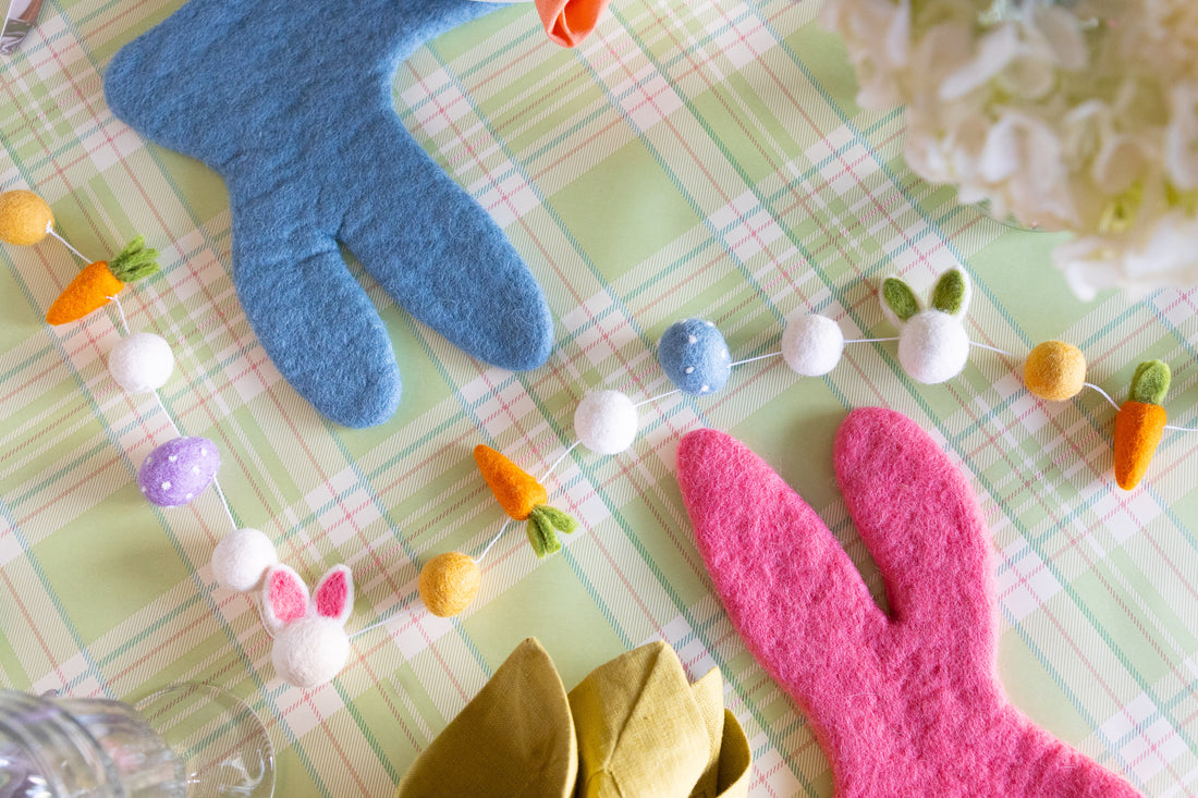 A close-up of an Easter themed table, featuring pink and blue felt bunny placemats, a felt garland, and a green plaid backdrop.