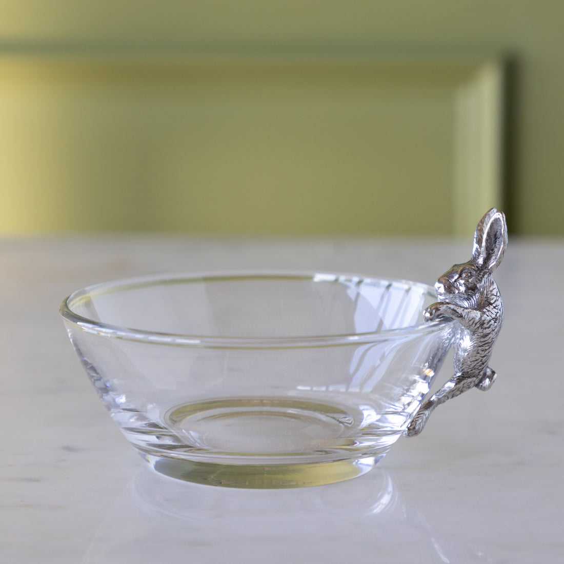 Clear glass bowl with a silver bunny figurine on the side, on a marble table.