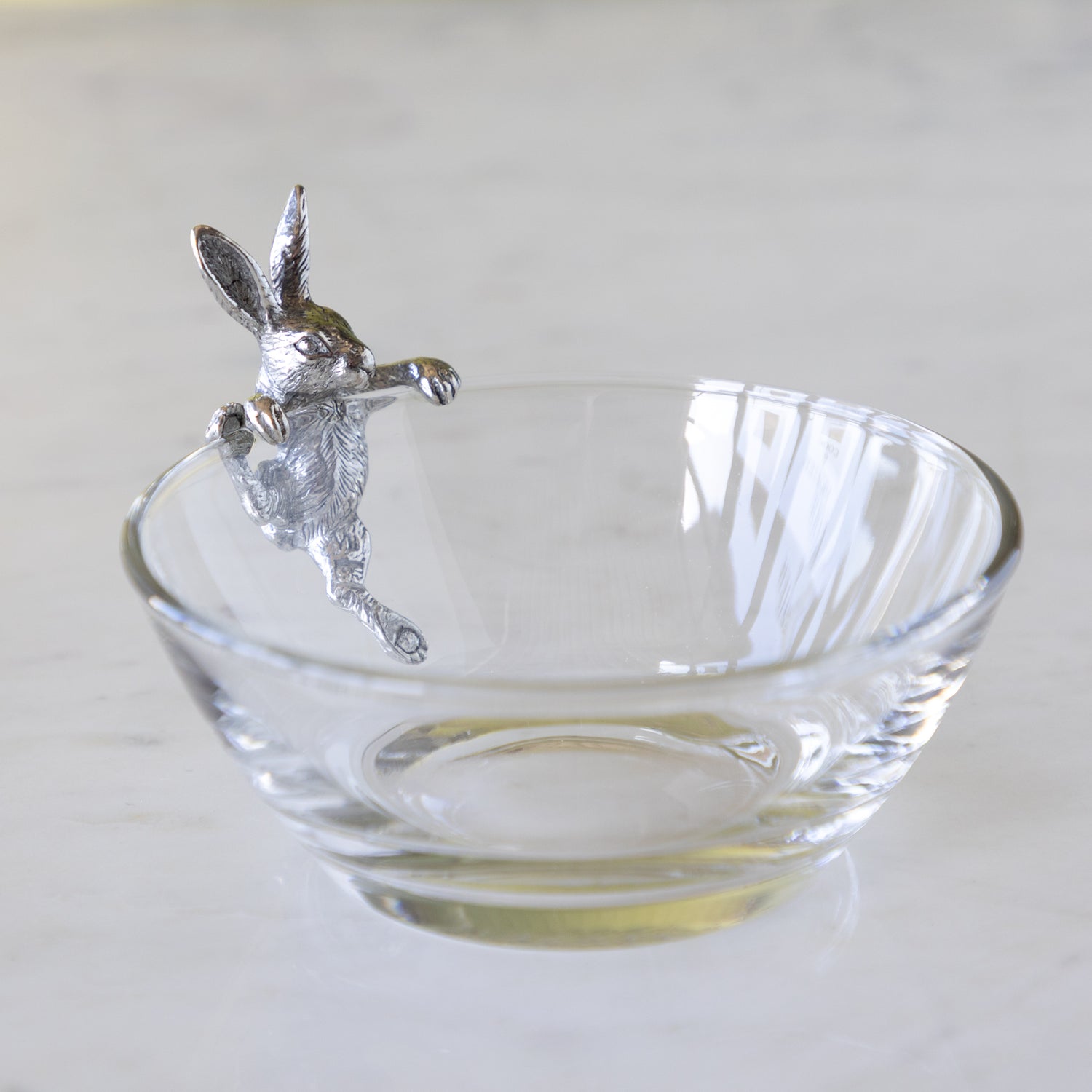 Clear glass bowl with a silver bunny figurine trying to climb into the bowl, on a white background.