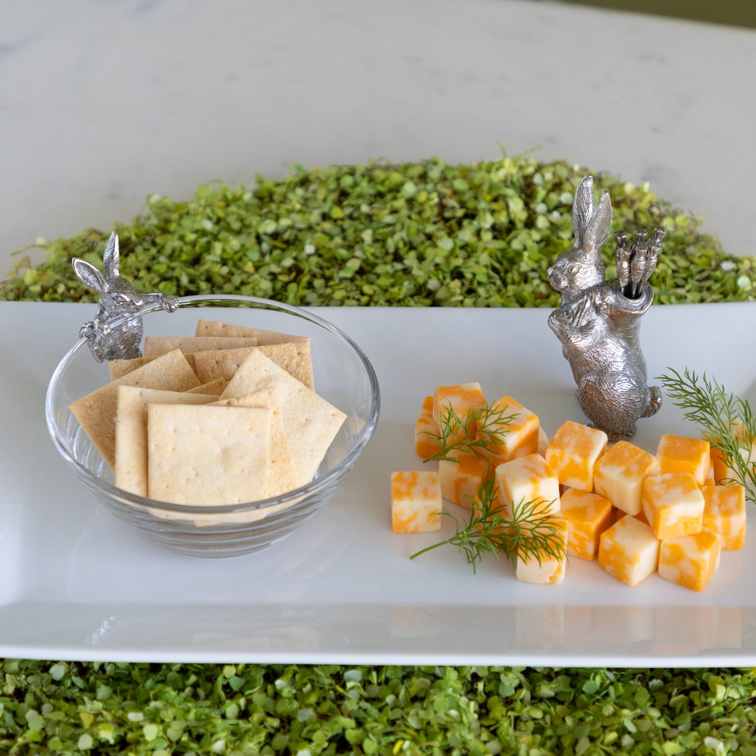 A serving platter with a Bunny Dip Bowl filled with crackers, cheese squares and a Rabbit with Carrot Picks on a moss placemat.