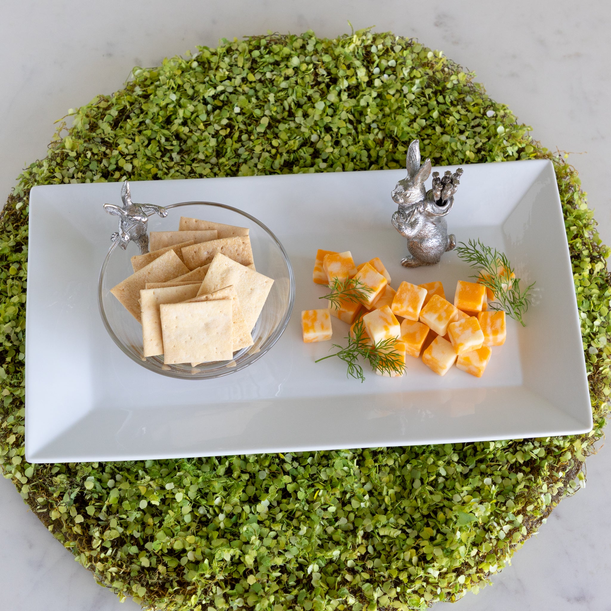 A serving platter with a Bunny Dip Bowl filled with crackers, cheese squares and a Rabbit with Carrots Picks on a moss placemat.