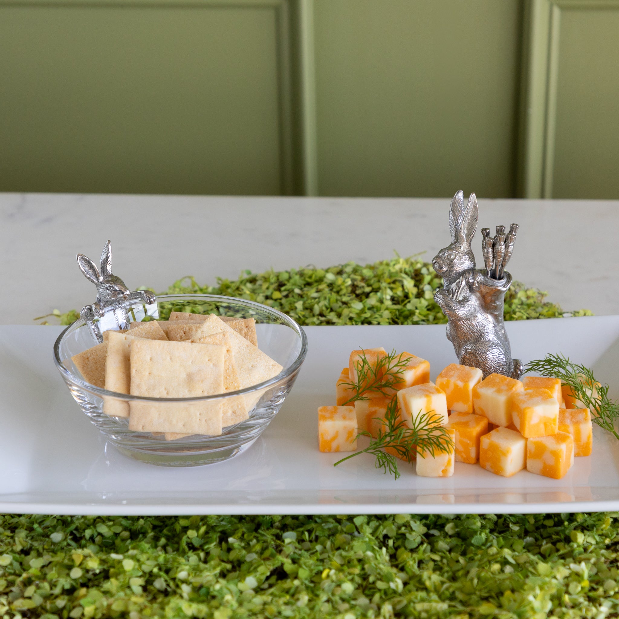 A serving platter with a Bunny Dip Bowl filled with crackers, cheese squares and a Rabbit with Carrots Picks on a moss placemat.