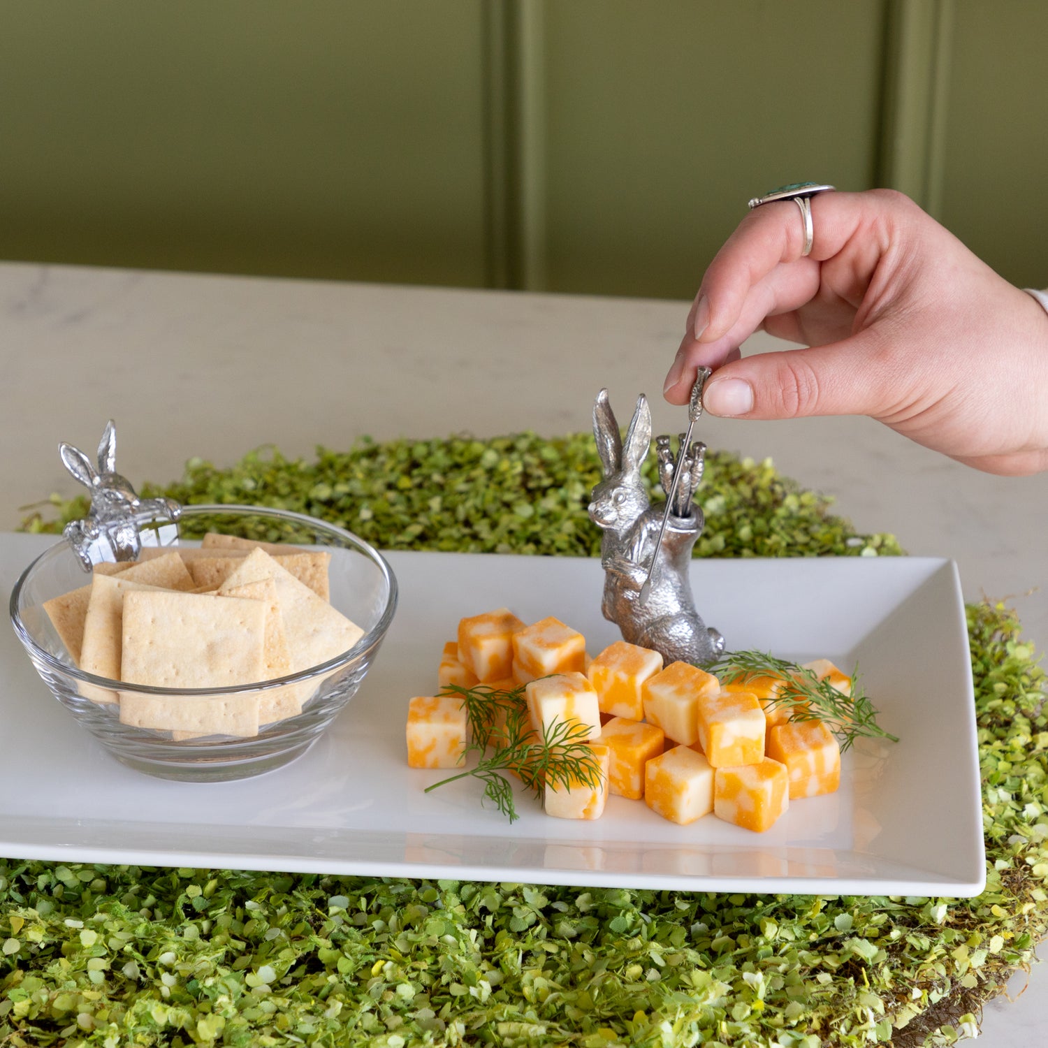 Person grabbing a carrot pick from a rabbit carrying picks, to serve cheese cubes from a platter.