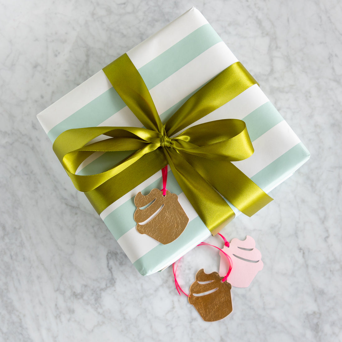 Gift wrapped in striped paper with a green ribbon and cupcake-shaped tags on a marble surface.