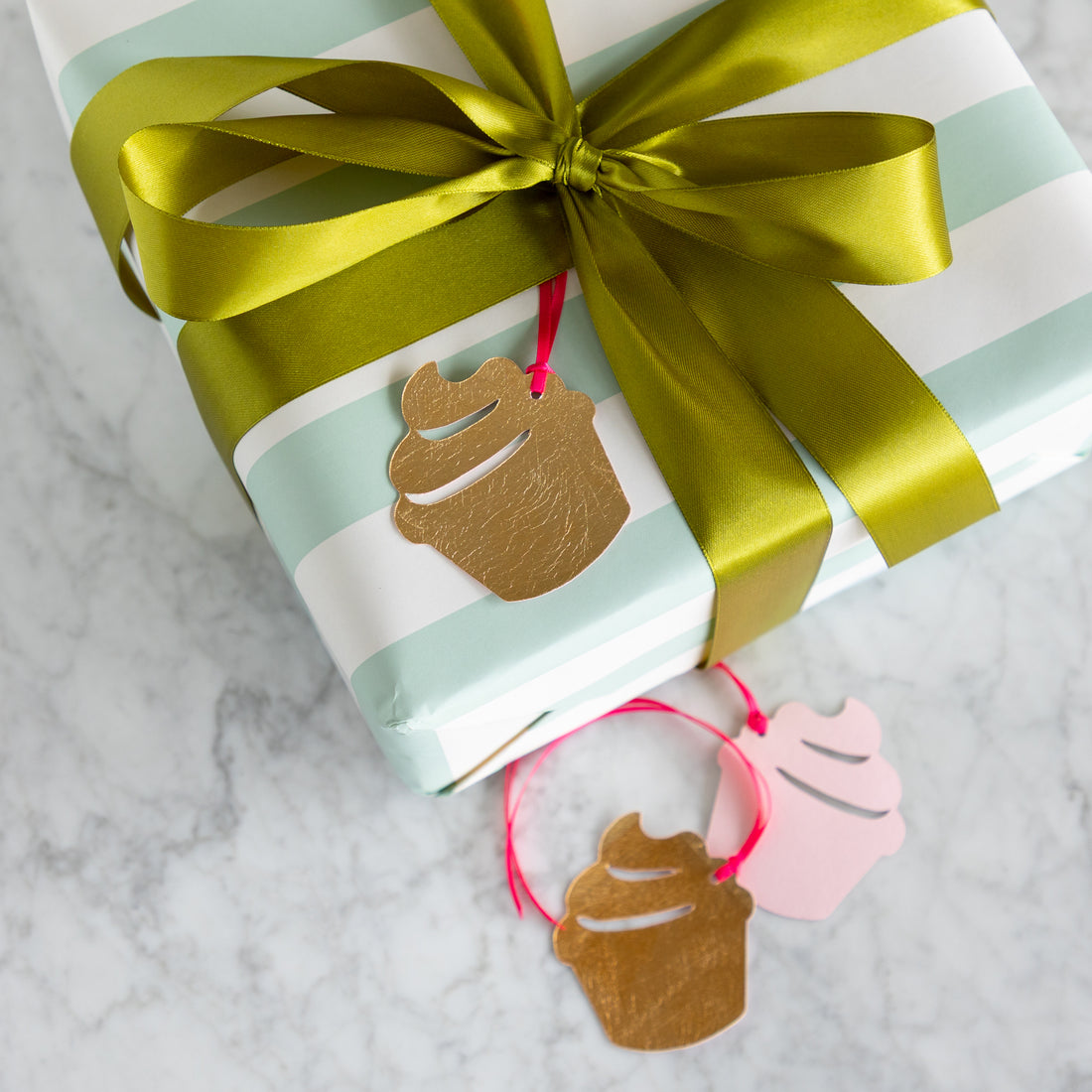 Gift wrapped in striped paper with a green ribbon and cupcake-shaped tags on a marble surface.