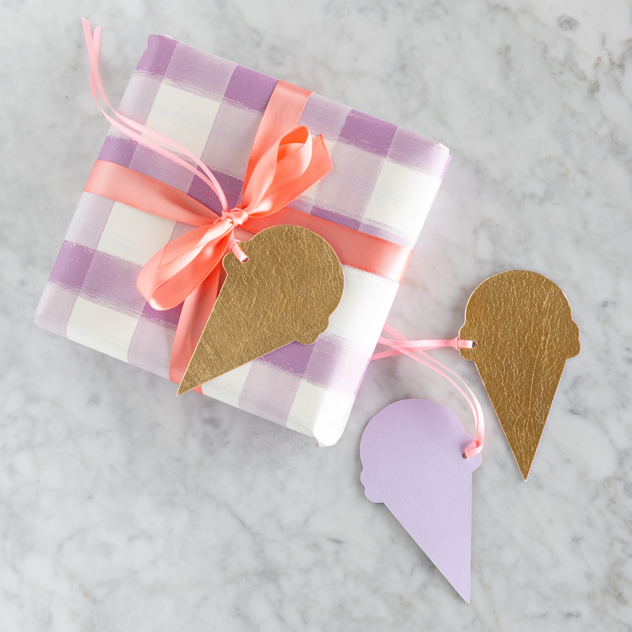 Gift wrapped in purple and white checkered paper with a pink ribbon, accompanied by reversible, gold-foiled and lavender ice cream cone-shaped tags on a marble surface.