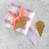 Gift wrapped in purple and white checkered paper with a pink ribbon, accompanied by reversible, gold-foiled and lavender ice cream cone-shaped tags on a marble surface.