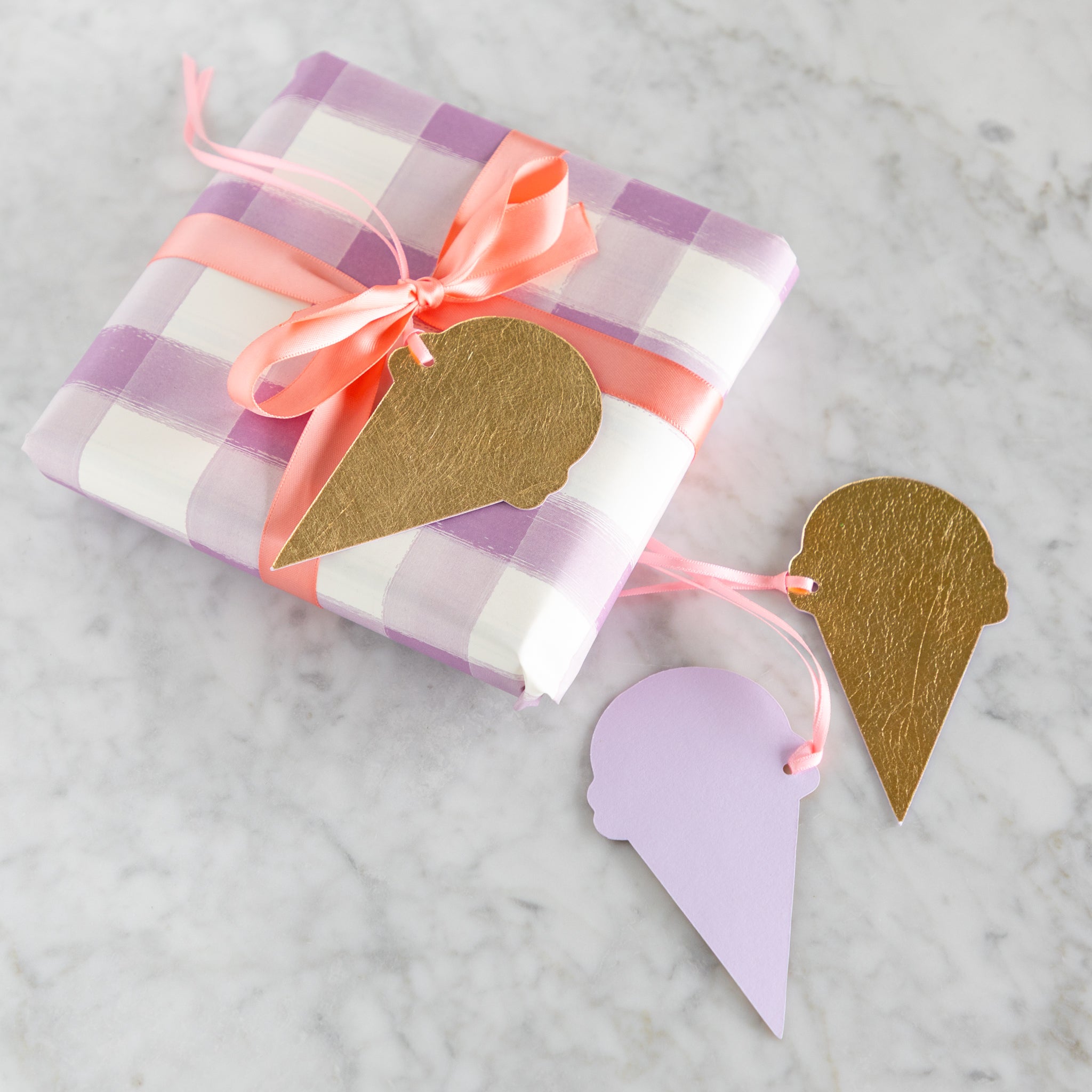 Gift wrapped in purple and white checkered paper with a pink ribbon, accompanied by reversible, gold-foiled and lavender ice cream cone-shaped tags on a marble surface.