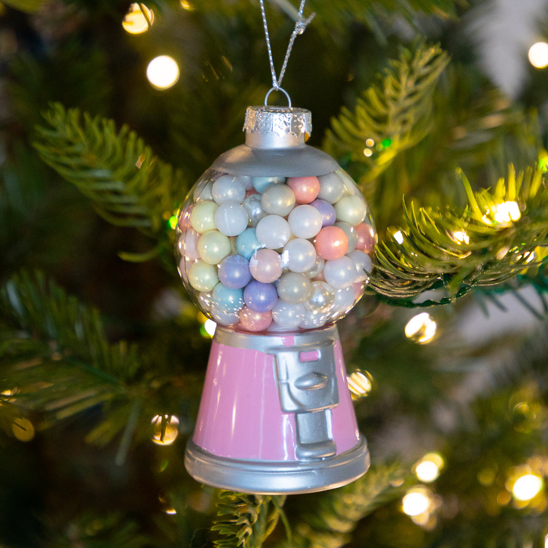 A gumball machine Christmas ornament hanging on a decorated Christmas tree. The ornament features a pink body, a silver metal base and top, and a clear dome filled with pastel-colored gumball beads.