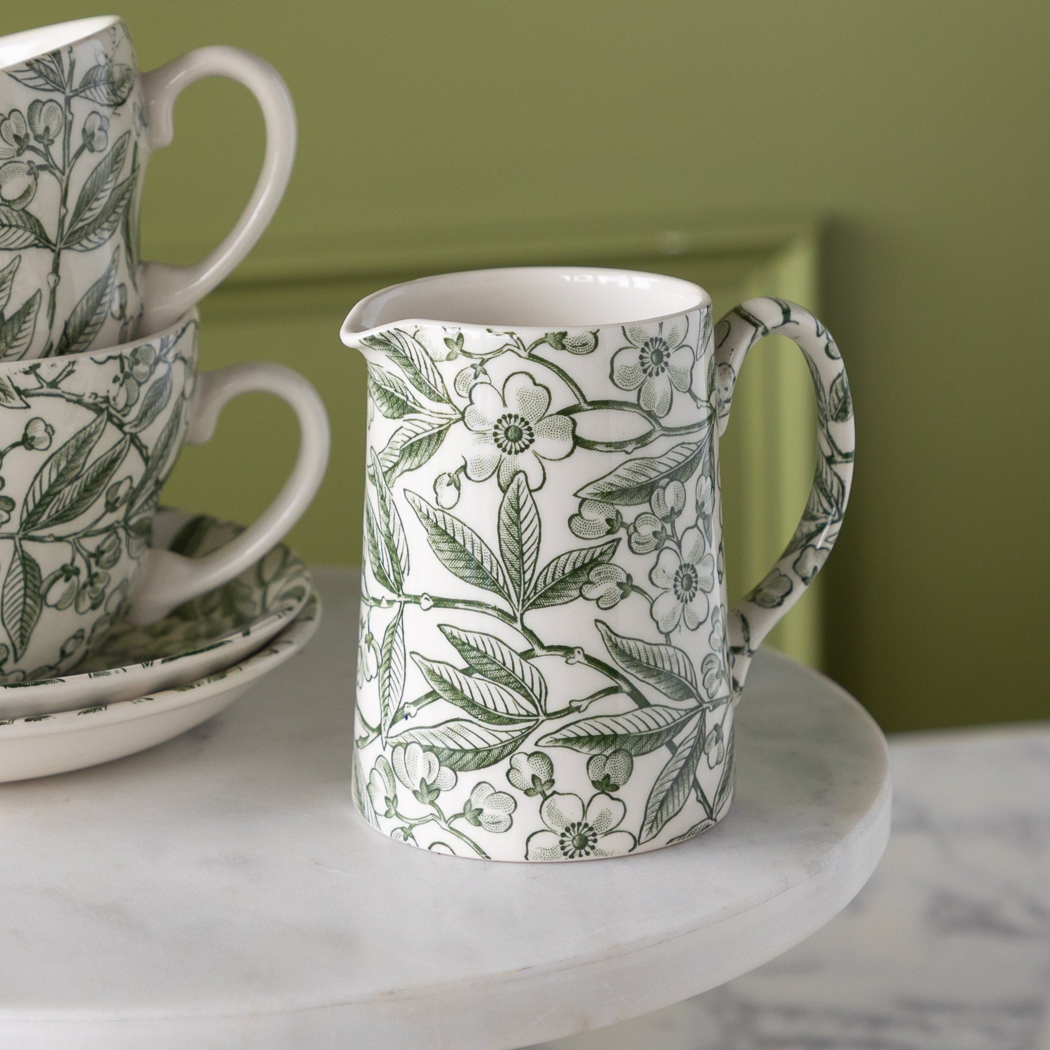 Burleigh Green Prunus Tankard Jug next to two cups and plates on a marble pedestal.