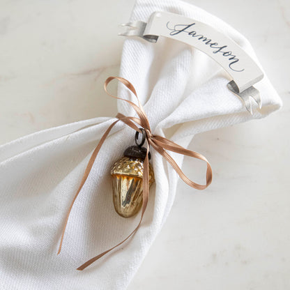 Glass acorn ornament tied around a napkin with a place card with the name &quot;Jameson&quot; written on it.