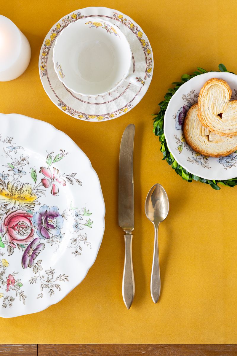 A table setting with floral plates, cups, and cutlery on a golden yellow felted table runner.