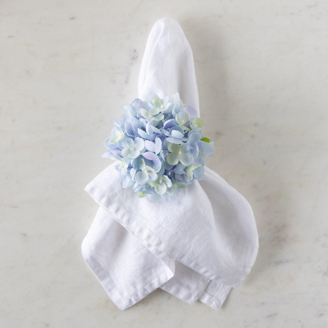White napkin with a blue Hydrangea Napkin Ring around it, on a marble surface.
