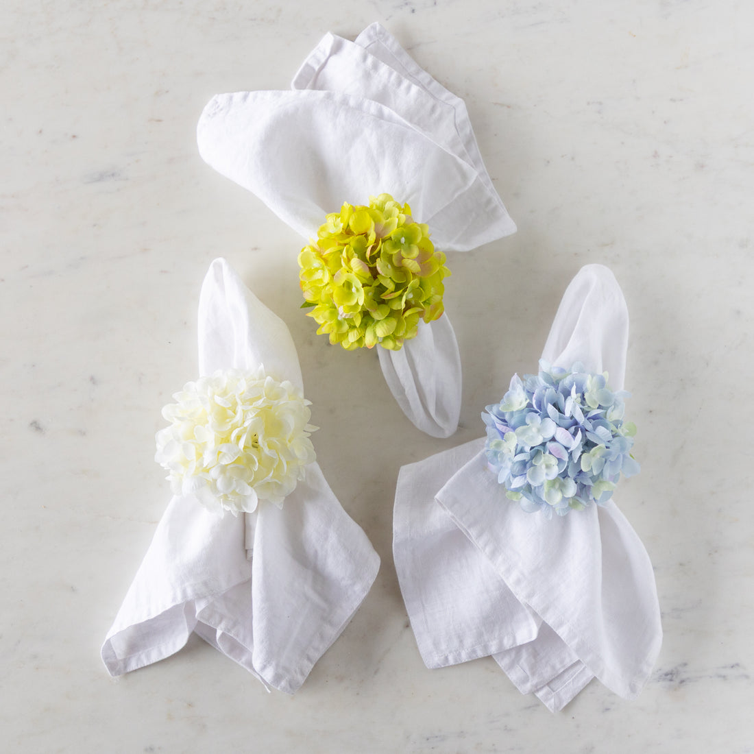 Three Hydrangea Napkin Rings in green, cream and blue, around white linen napkins.