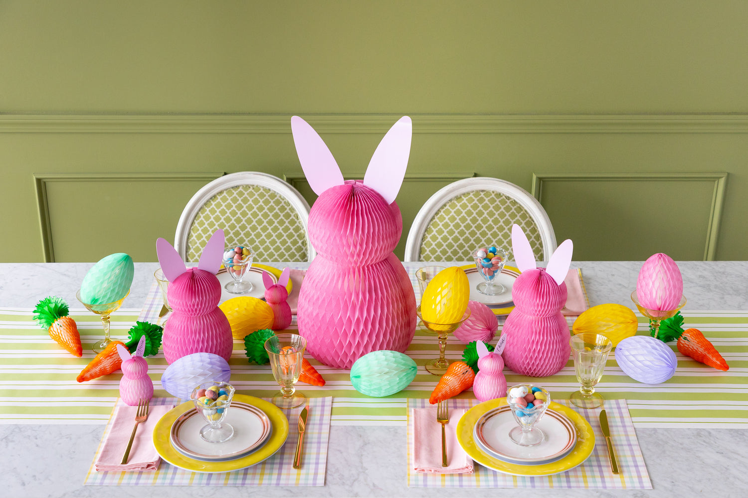 Easter-themed table setting with pink bunny decorations, colorful eggs, and carrots on a striped tablecloth.