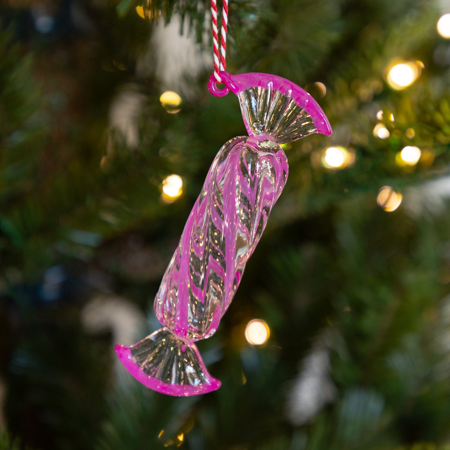 Long, dark pink glass candy ornament hanging on a Christmas tree with blurred lights in the background.