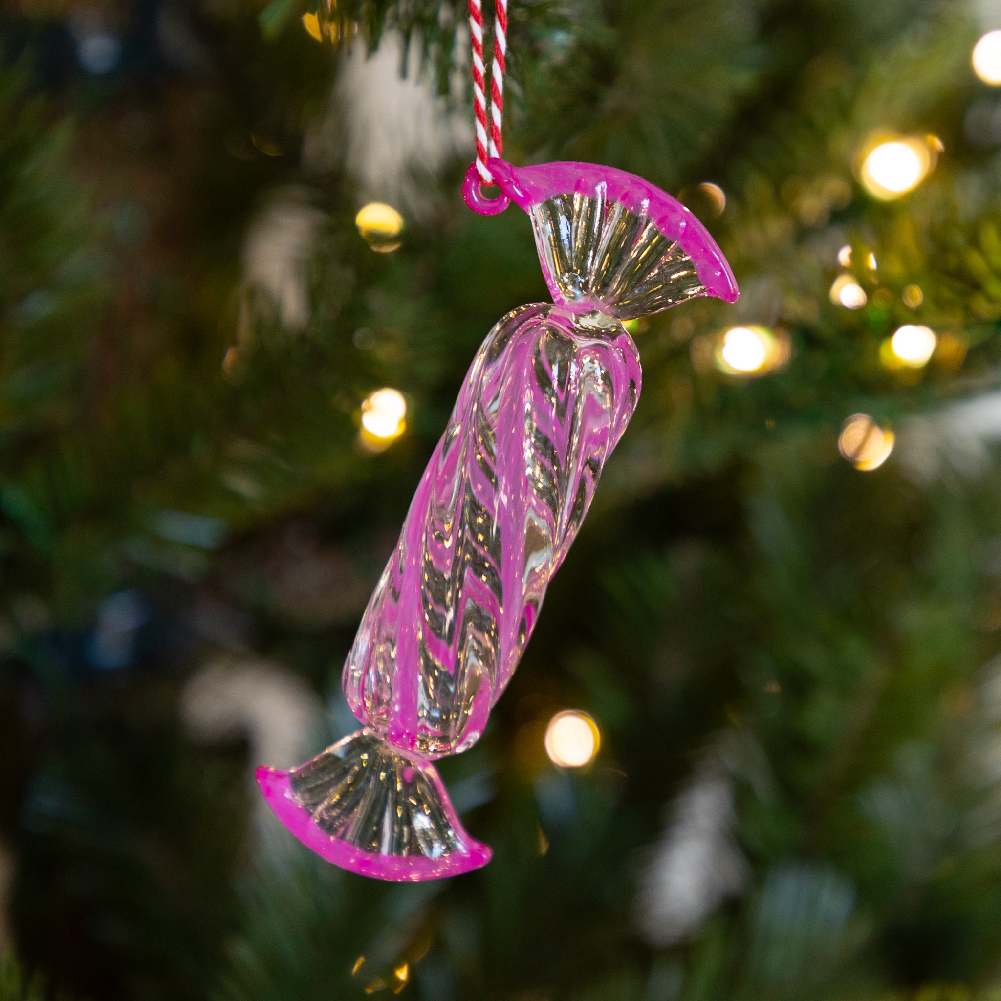 Long, dark pink glass candy ornament hanging on a Christmas tree with blurred lights in the background.