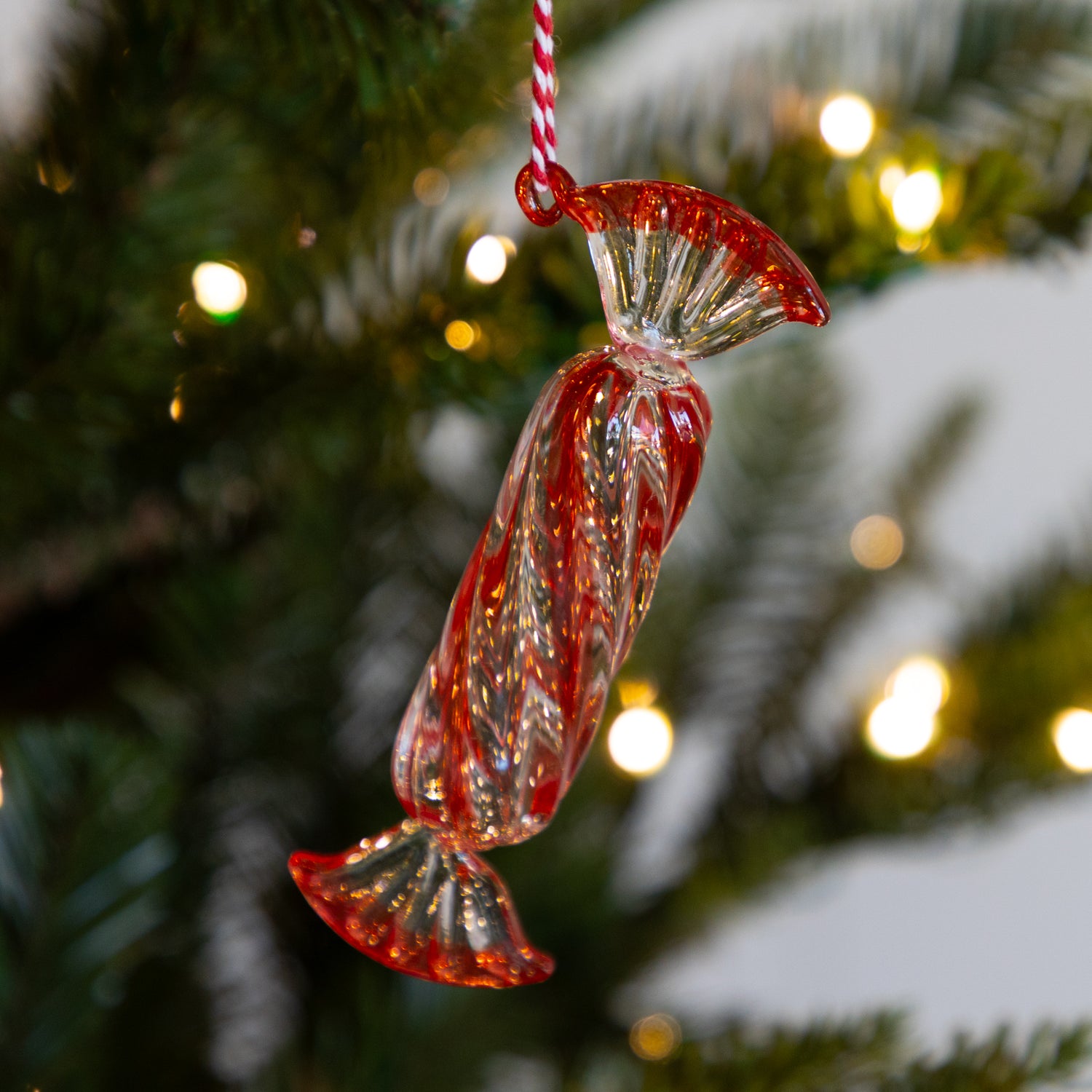 Long, red glass candy ornament hanging on a Christmas tree with blurred lights in the background.