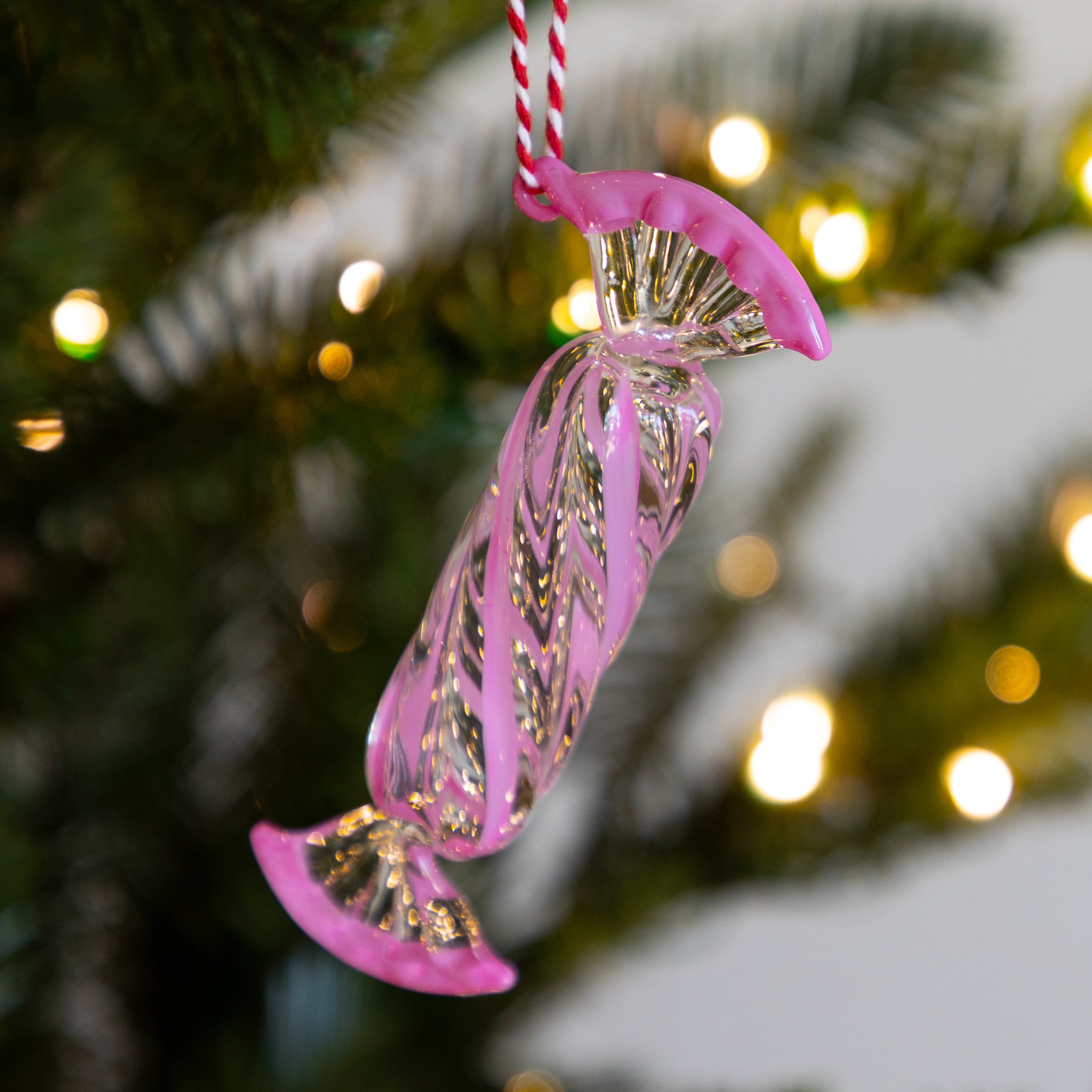Long, pink glass candy ornament hanging on a Christmas tree with blurred lights in the background.